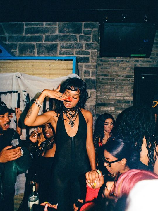 Colour flash photograph in a crowded bar: a woman in a black halter dress shields her eyes with her hand while people dance around her