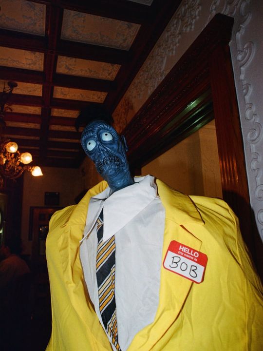 Colour photograph in a wood-panelled room: a figure in a blue monster mask and a bright yellow suit with a name tag reading Hello my name is Bob